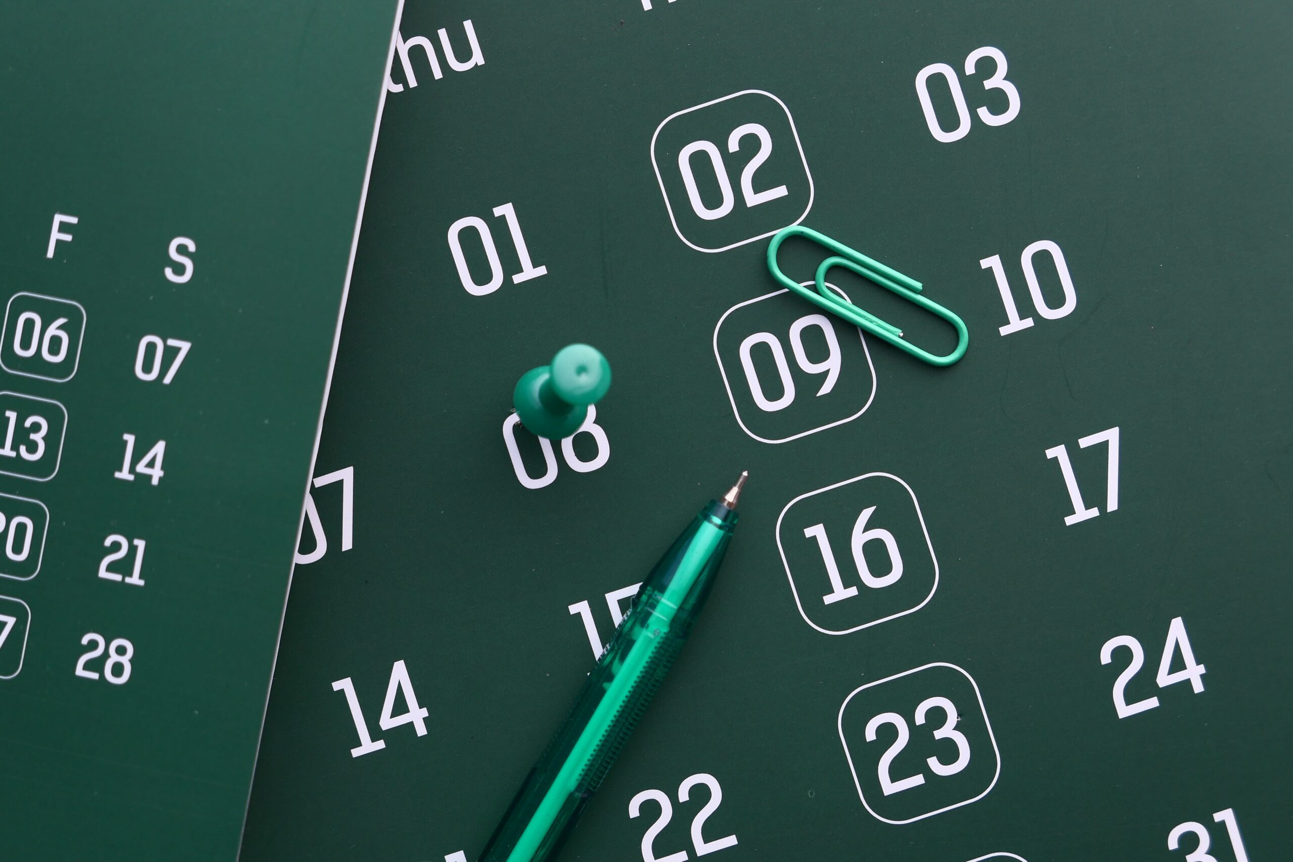 Green wall calendar with a green pin, a paper clip and a pen lying across the dates