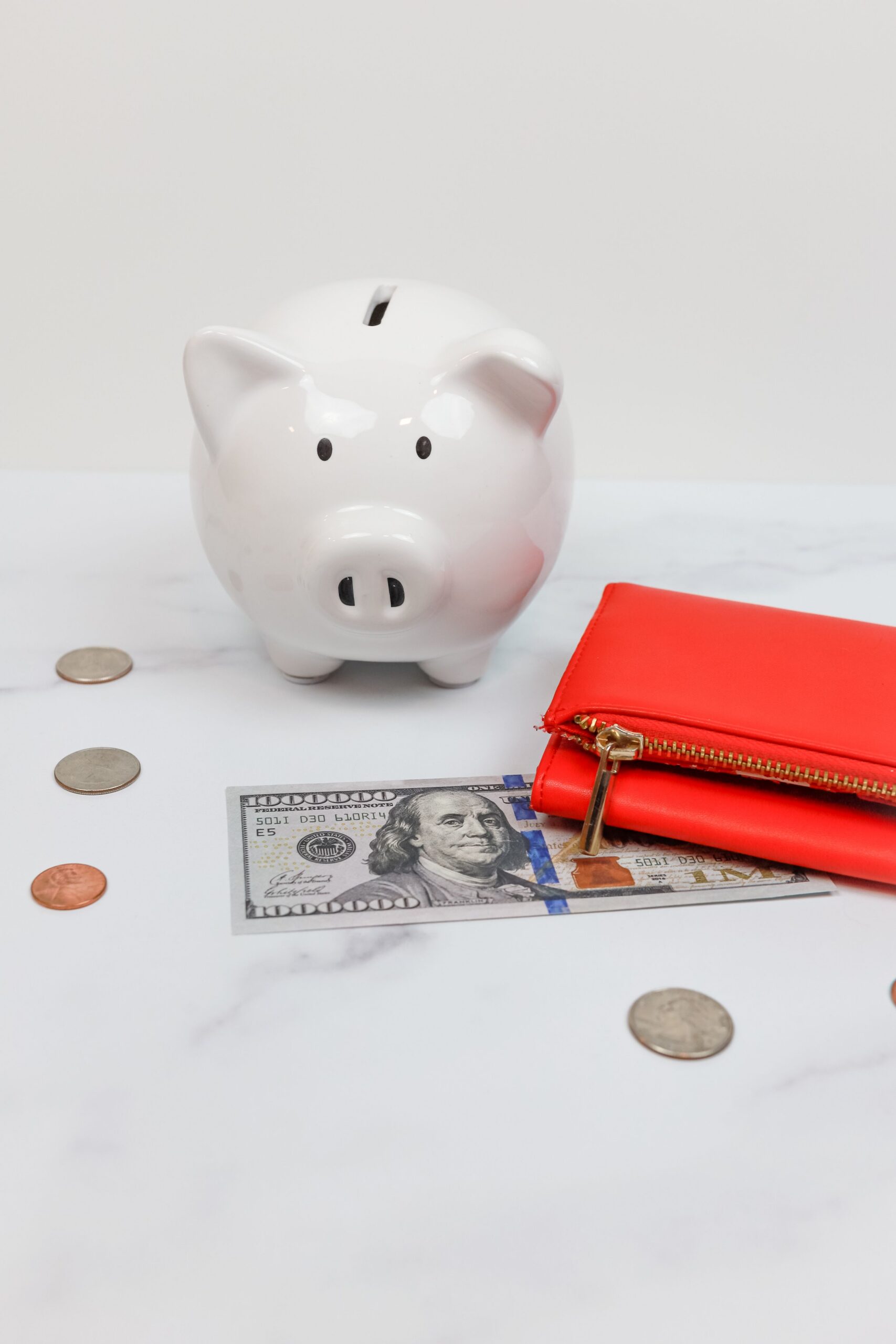 White piggy bank beside a bill, loose coins and a red wallet on a marble surface