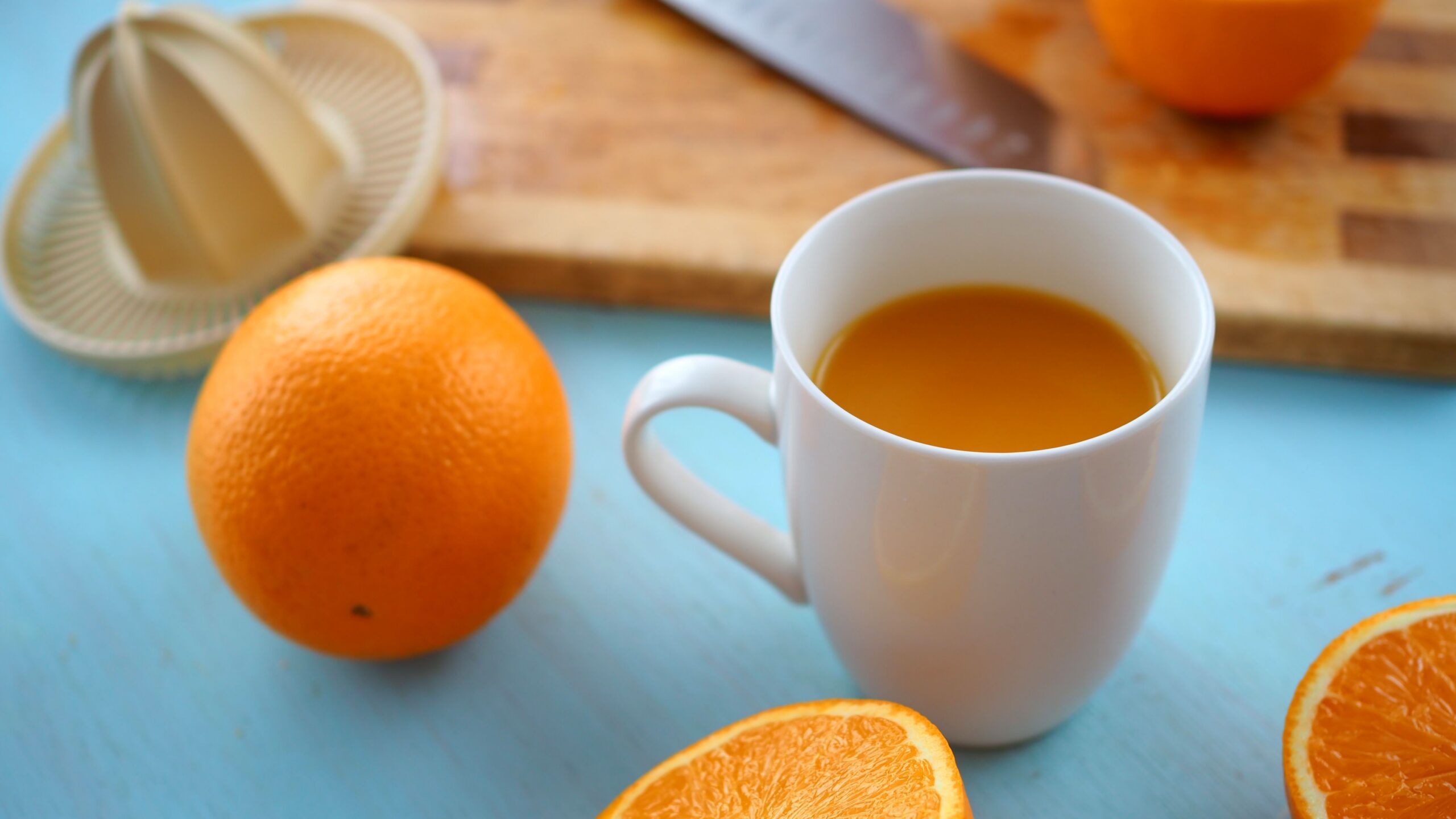 Orange juice in a white cup beside whole and halved oranges and a hand juicer