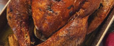A roasted turkey with a deep brown skin in a rectangular baking pan, seen from above, with green beans and mashed potatoes beside it