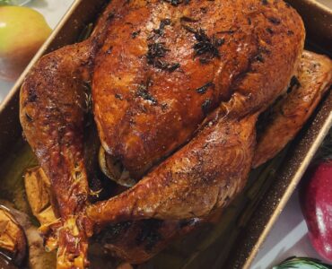A roasted turkey with a deep brown skin in a rectangular baking pan, seen from above, with green beans and mashed potatoes beside it