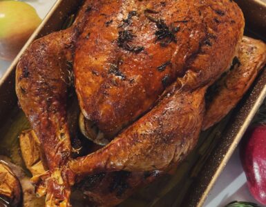 A roasted turkey with a deep brown skin in a rectangular baking pan, seen from above, with green beans and mashed potatoes beside it