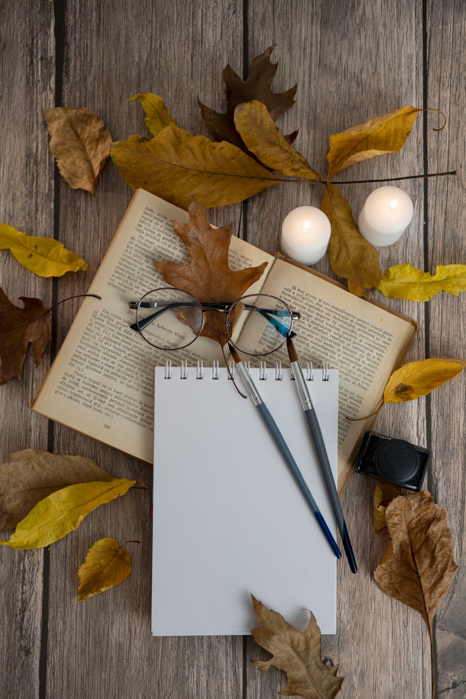 Yellow and brown autumn leaves around an open book, round glasses, a blank notepad and two candles on a wooden table