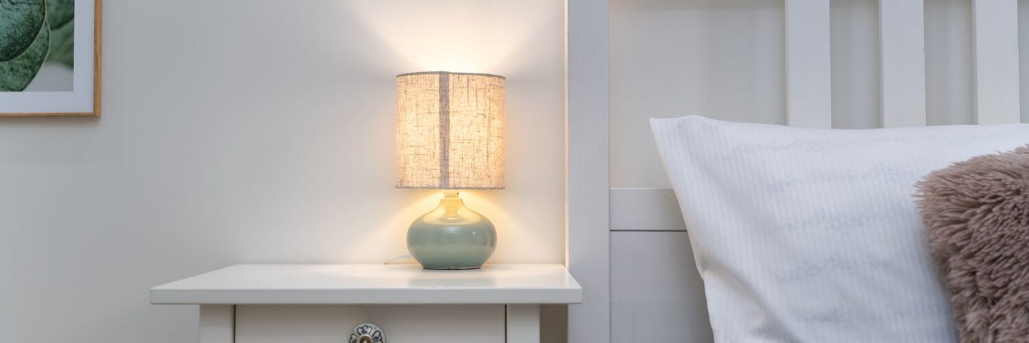 A small lit table lamp on a white nightstand beside a bed with a gray cover and white pillow