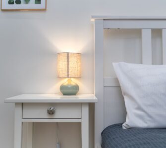 A small lit table lamp on a white nightstand beside a bed with a gray cover and white pillow
