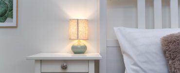A small lit table lamp on a white nightstand beside a bed with a gray cover and white pillow