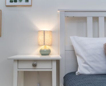 A small lit table lamp on a white nightstand beside a bed with a gray cover and white pillow