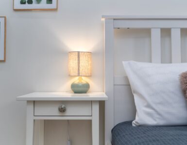 A small lit table lamp on a white nightstand beside a bed with a gray cover and white pillow