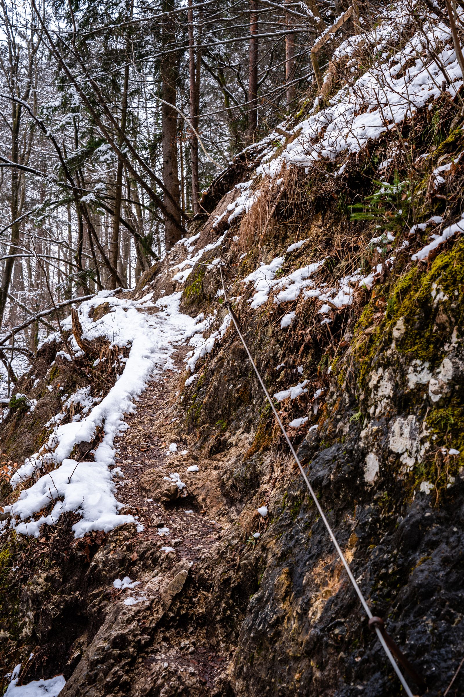 Narrow dirt trail along a rock wall with patches of snow and a cable railing