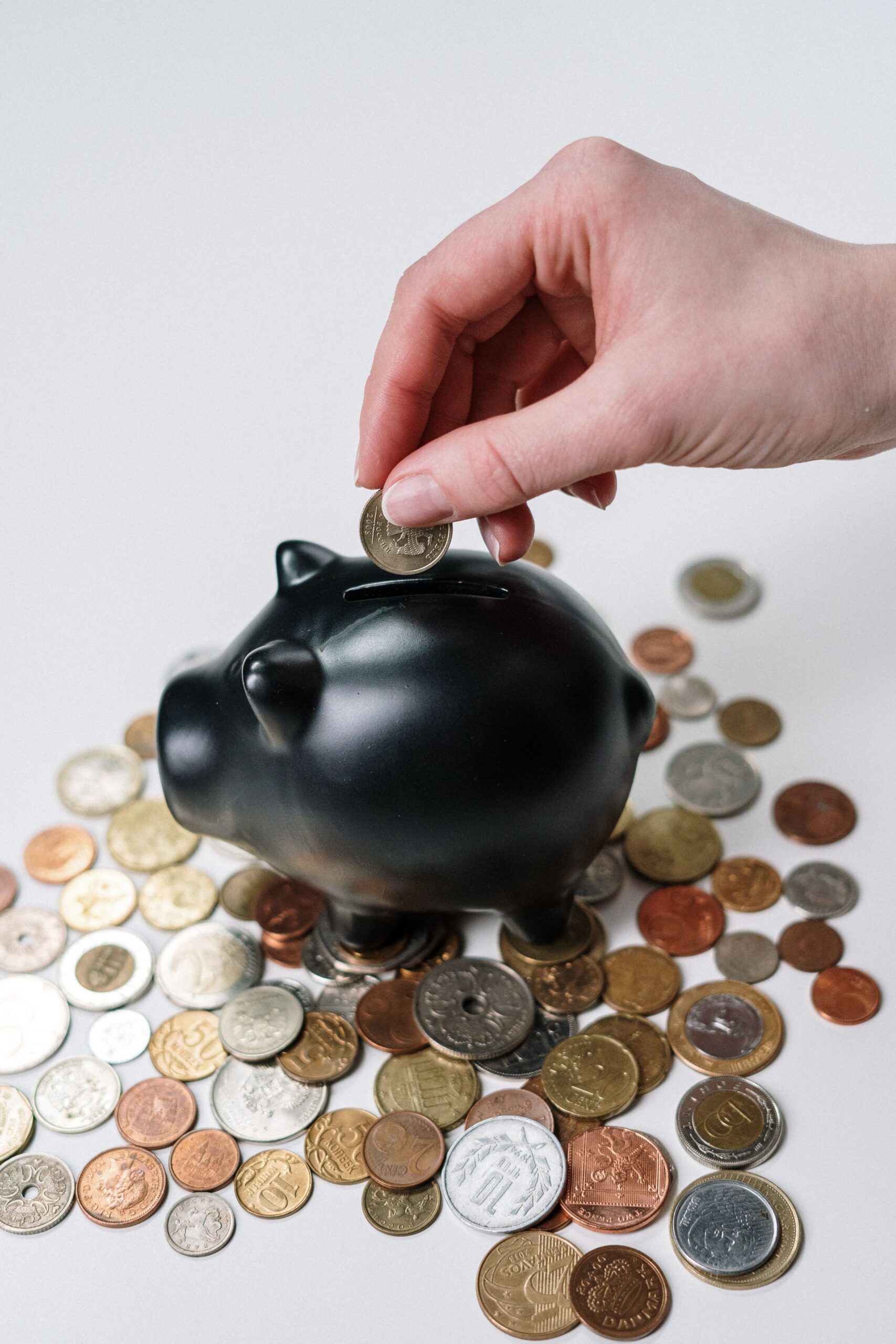 A hand dropping a coin into a black piggy bank surrounded by loose coins on a white surface