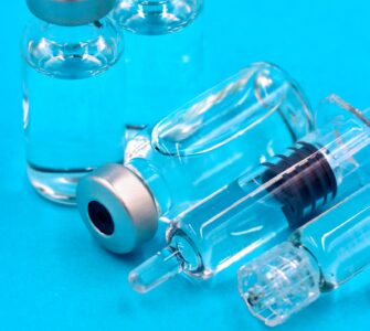 Two clear glass vials with silver caps and a prefilled syringe lying on a bright blue surface