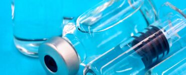 Two clear glass vials with silver caps and a prefilled syringe lying on a bright blue surface