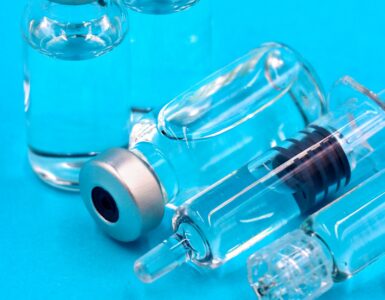Two clear glass vials with silver caps and a prefilled syringe lying on a bright blue surface