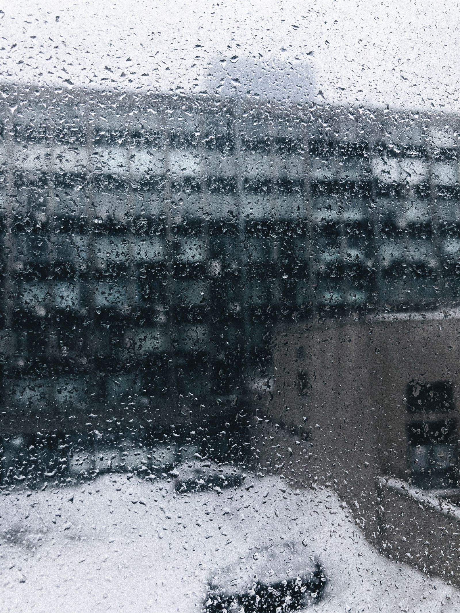 Water droplets covering a window pane, with a snow-covered roof and buildings blurred beyond