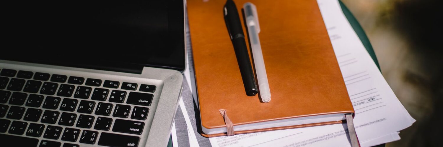 A tan notebook with two pens resting on stacked papers beside a laptop