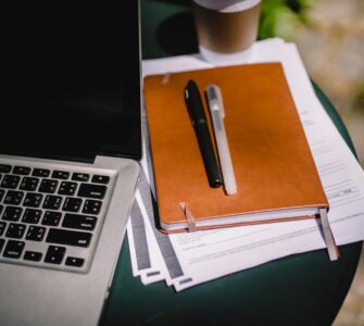 A tan notebook with two pens resting on stacked papers beside a laptop