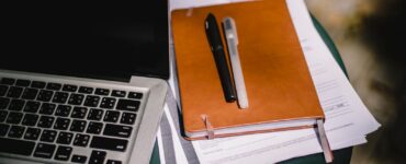 A tan notebook with two pens resting on stacked papers beside a laptop