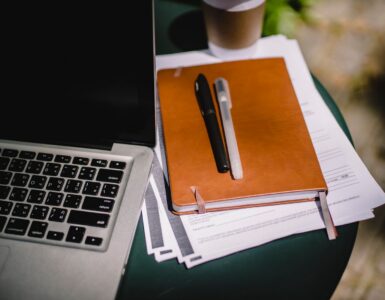 A tan notebook with two pens resting on stacked papers beside a laptop