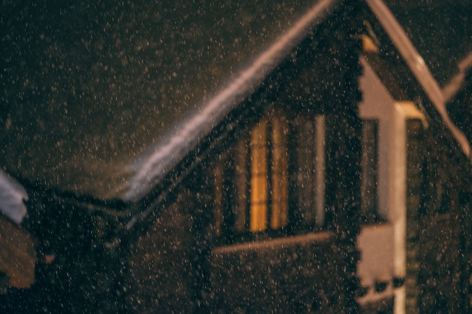 A small wooden cottage at night with snow falling and one window glowing orange