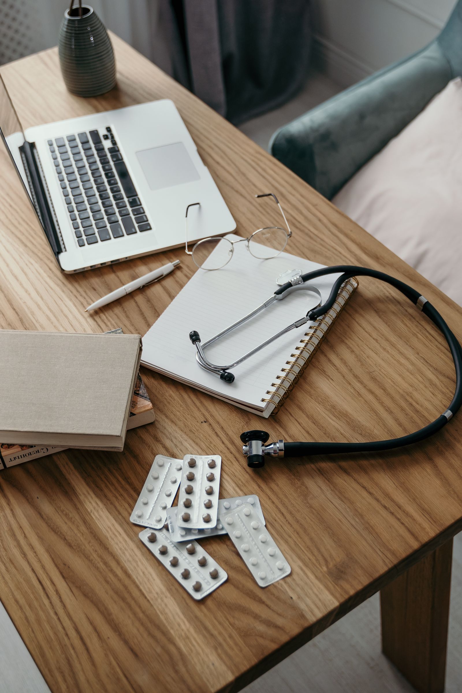A stethoscope, pill blister packs, a notebook and a laptop on a wooden desk