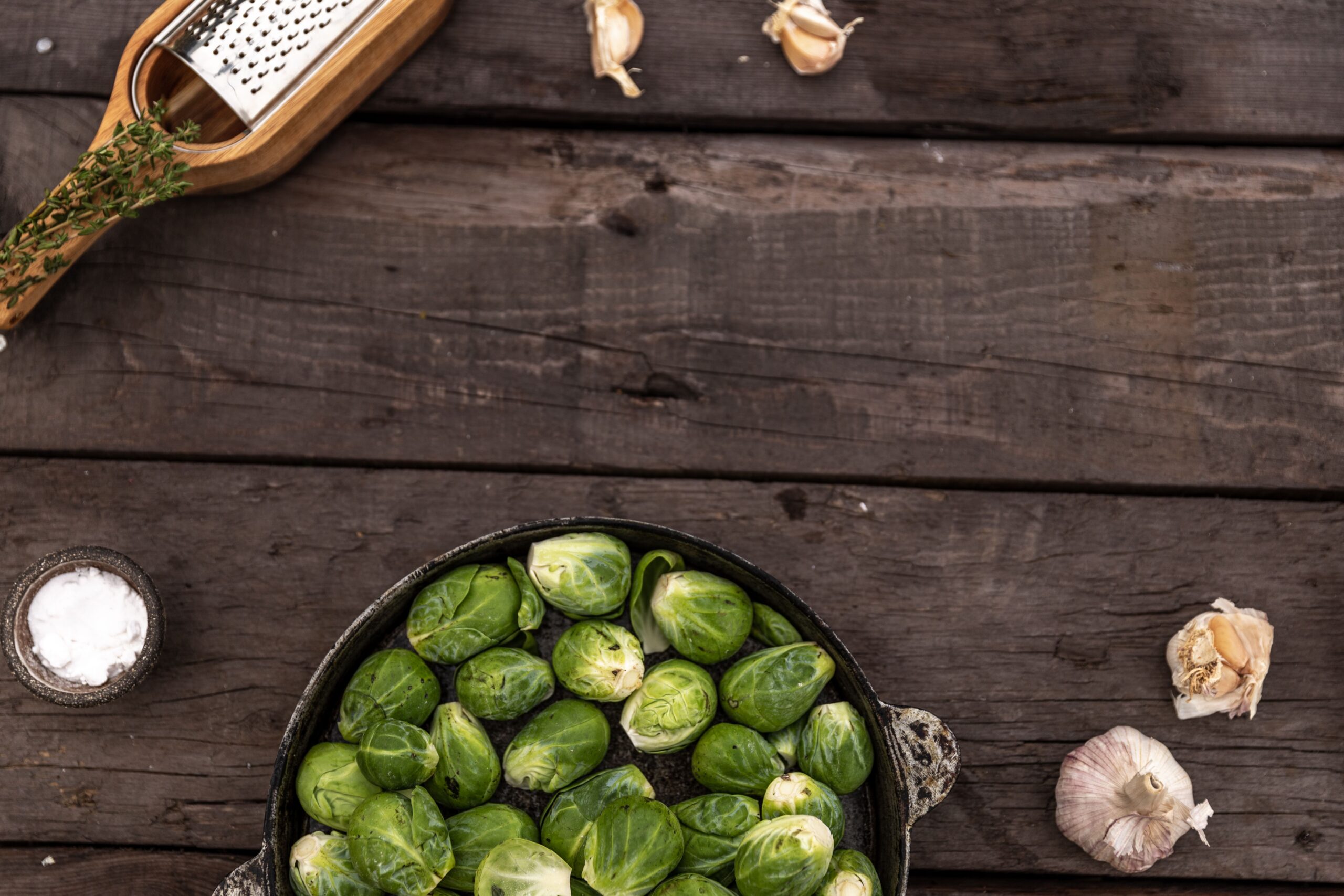 Raw Brussels sprouts in a cast-iron pan on a wooden table with garlic and a grater