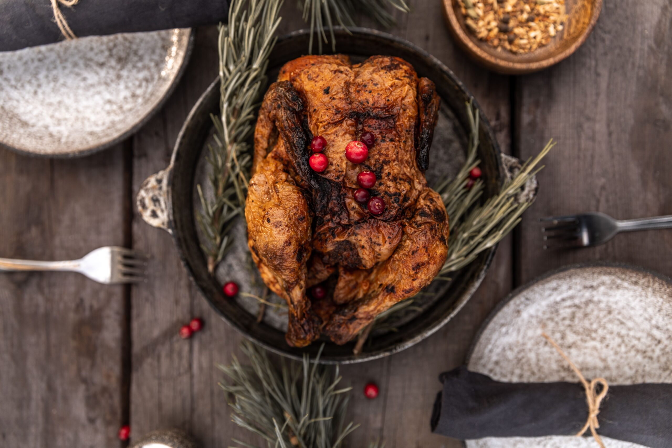 A whole roasted bird in a dark pan with rosemary and cranberries on a wooden table