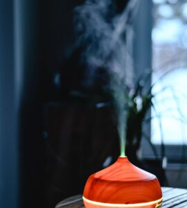 A small tabletop mist unit glowing on a wooden table beside a window, releasing a thin plume of vapor