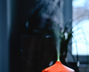 A small tabletop mist unit glowing on a wooden table beside a window, releasing a thin plume of vapor