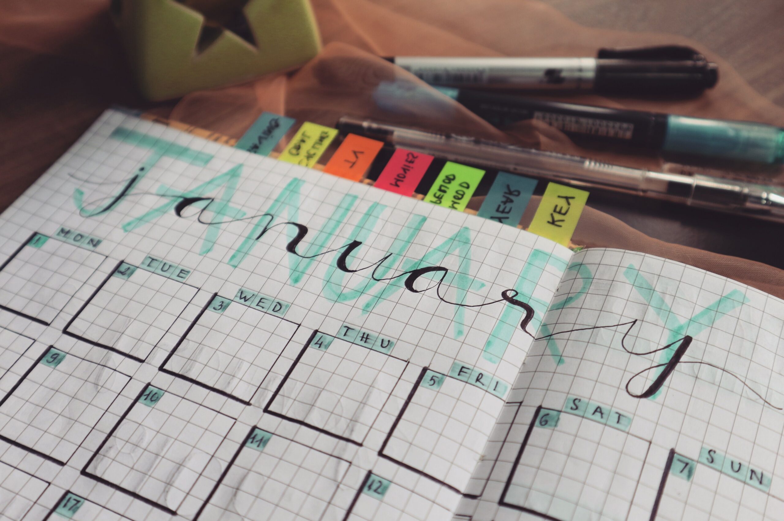 A hand-lettered January calendar page in a grid notebook with colored sticky tabs and pens