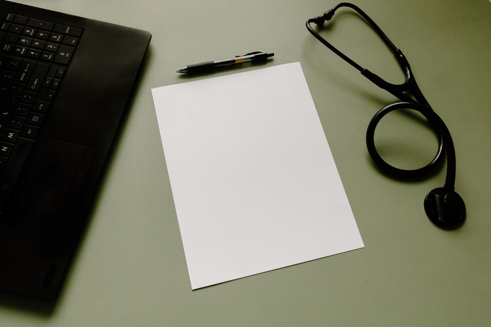A blank sheet of paper, a pen, a laptop and a stethoscope on a green desk