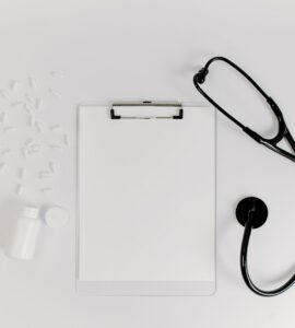 A blank clipboard, a stethoscope and scattered white capsules on a white desk