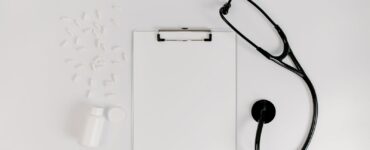 A blank clipboard, a stethoscope and scattered white capsules on a white desk