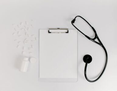 A blank clipboard, a stethoscope and scattered white capsules on a white desk