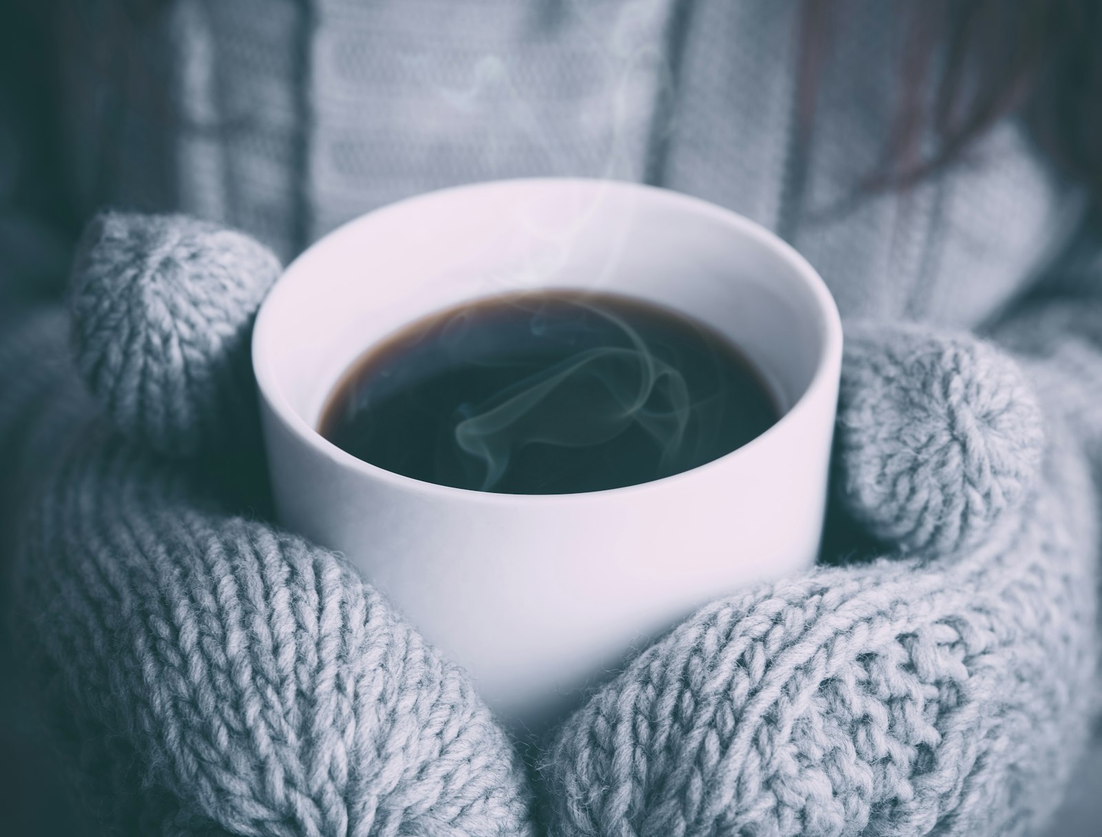 Hands in gray knit mittens holding a white mug of steaming black coffee