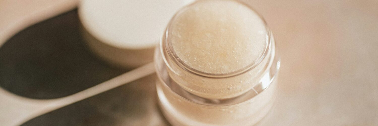 A small clear jar of pale lip care product with its lid set beside it on a marble surface