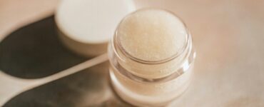 A small clear jar of pale lip care product with its lid set beside it on a marble surface