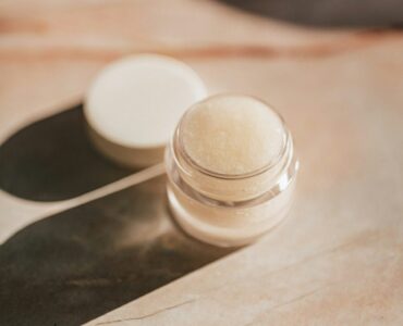 A small clear jar of pale lip care product with its lid set beside it on a marble surface
