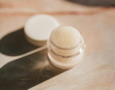 A small clear jar of pale lip care product with its lid set beside it on a marble surface