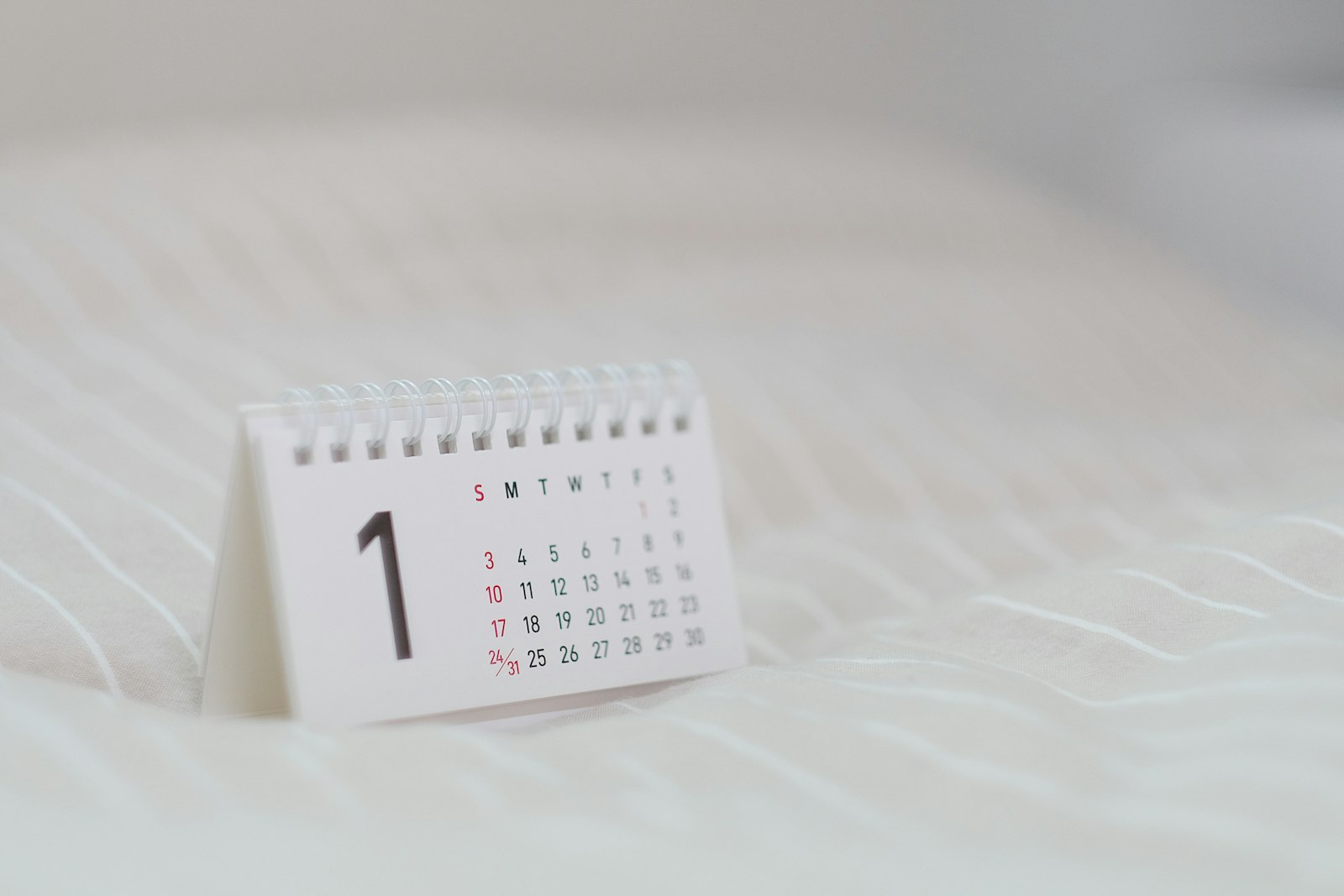 A small spiral desk calendar standing on white fabric, open to a month page