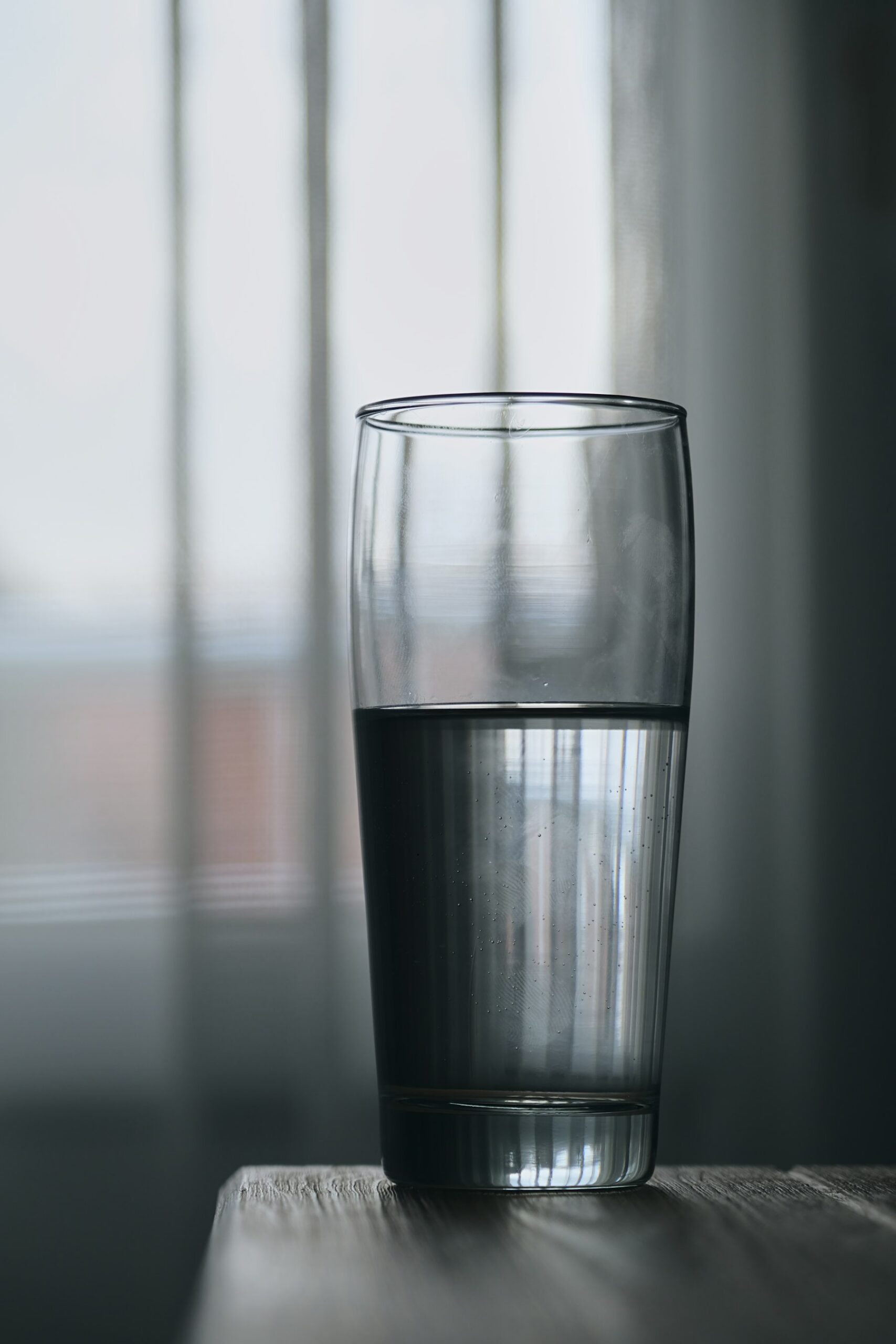 A drinking glass about half full of water on a table in front of a window