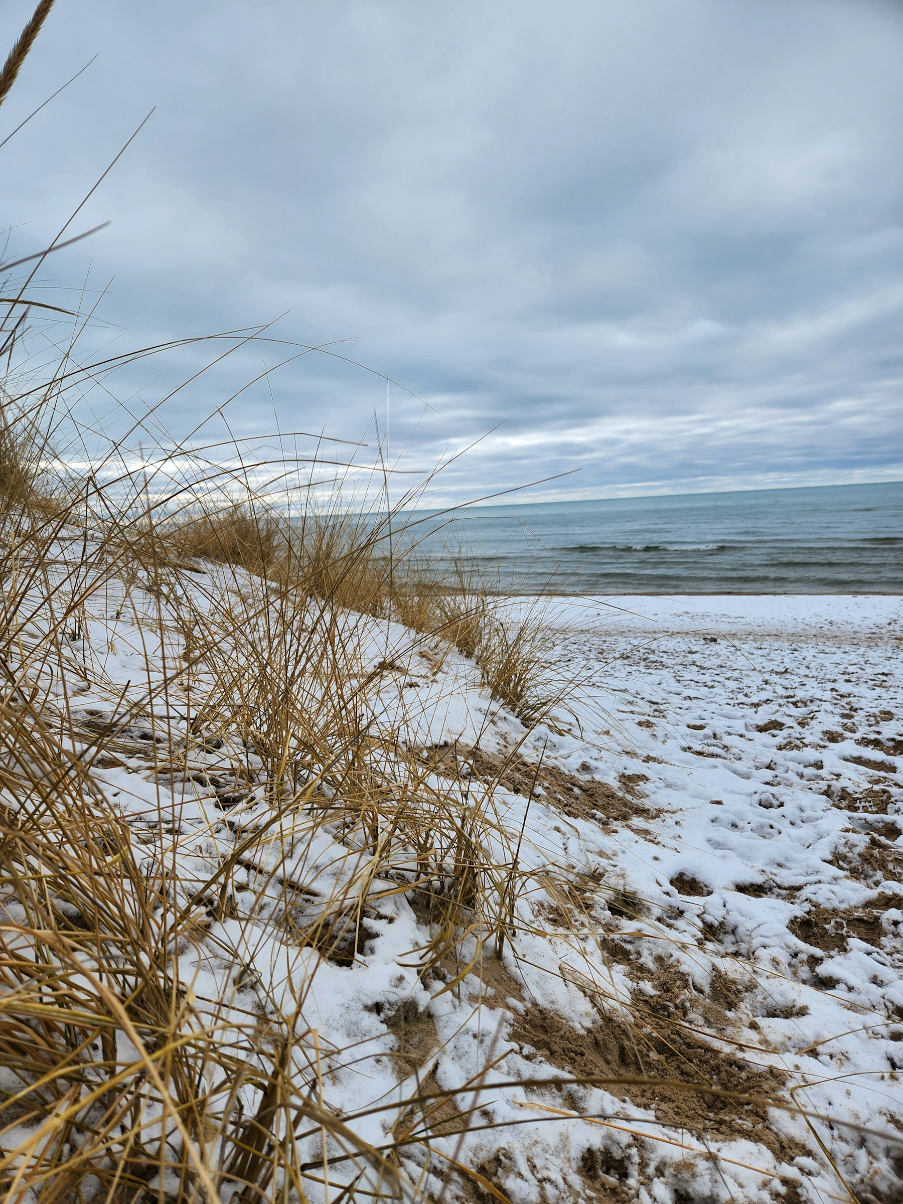 A sandy beach dusted with snow beside open water under a gray sky